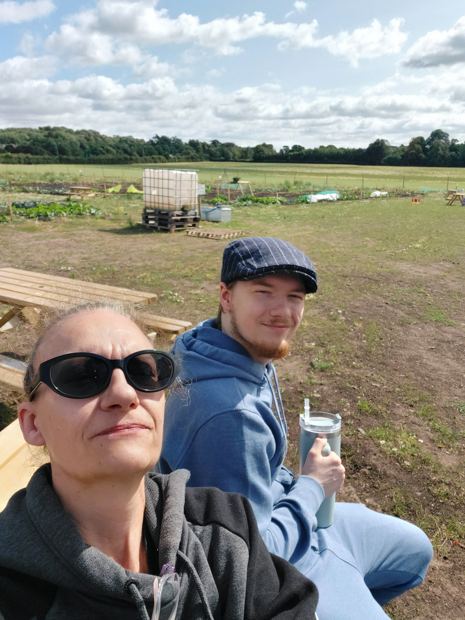 Two people sit outdoors in a field, with grassy areas and clouds in the background.
